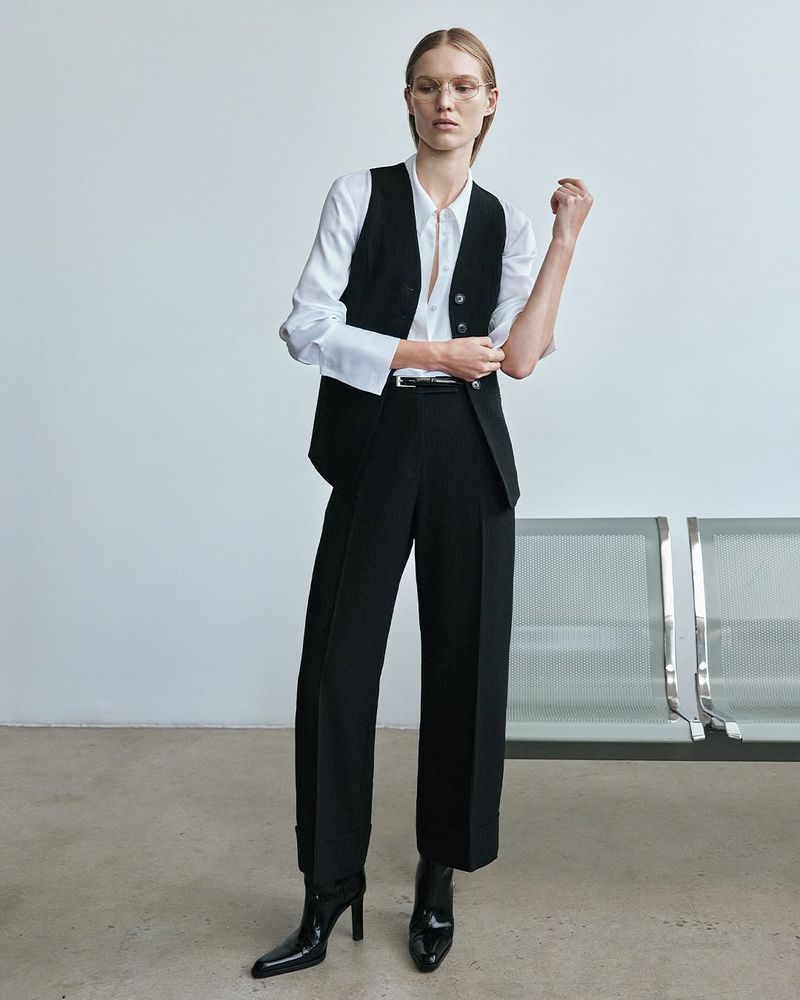 Full-length shot of a woman in a white blouse, black vest, and wide-leg trousers, standing in front of a white wall and metal bench.