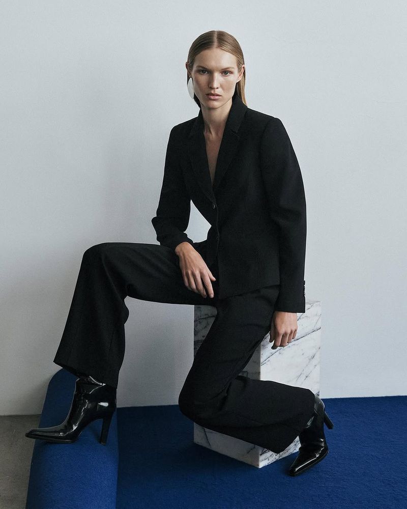 A fashionable woman in a black pantsuit poses indoors, seated on a marble block, showcasing modern professional attire and high-heeled boots.