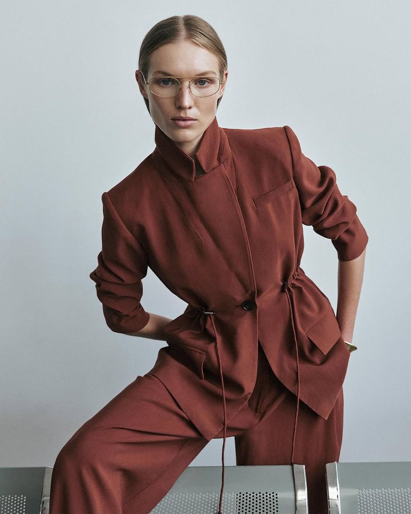 Woman poses in a rust-colored suit and eyeglasses against a neutral background, with an eye-level medium shot capturing fashion and style.