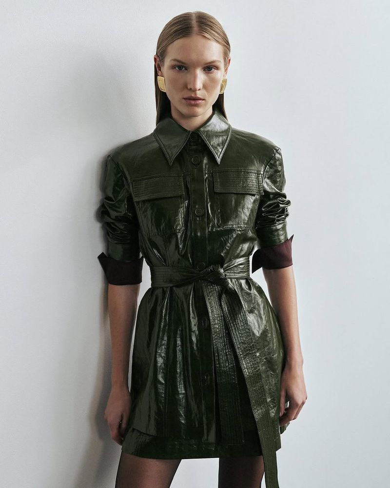 A fair-skinned adult female poses indoors wearing a shiny, green collared dress with a tie-belt, gold earrings, and dark stockings against a white wall.