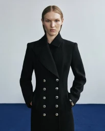 Model wearing a black double-breasted coat with silver buttons against a plain light wall and blue floor