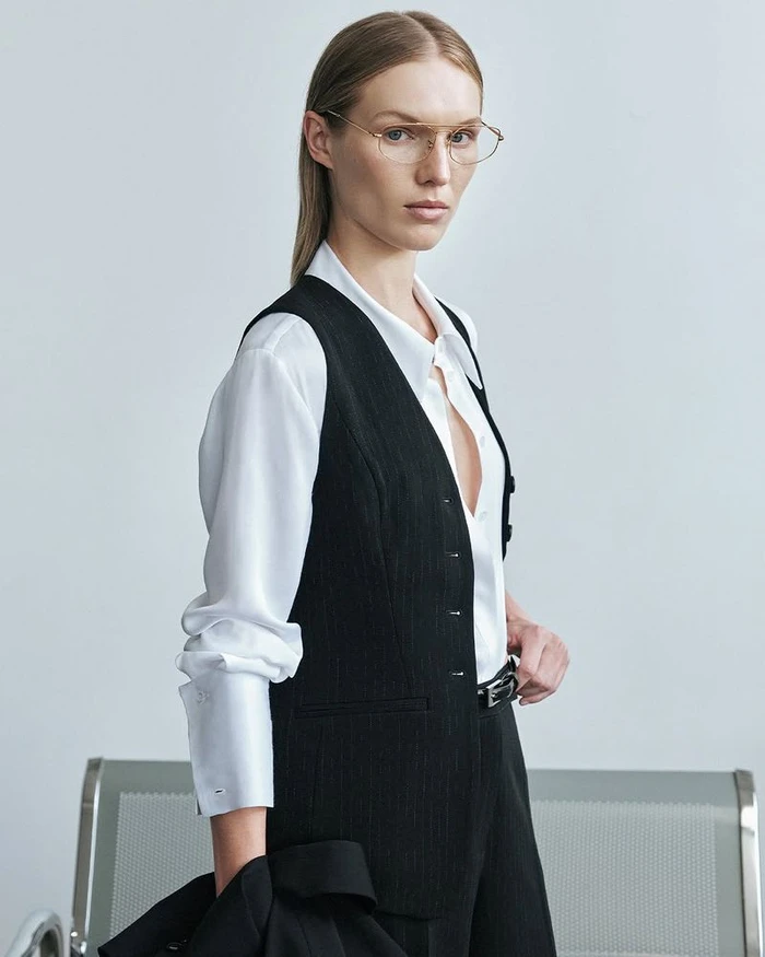Model wearing a white shirt and black vest with eyeglasses against a light grey studio background.