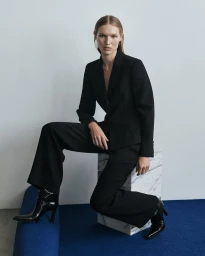 Model in black suit and patent boots sitting on a marble block for Cue July 25