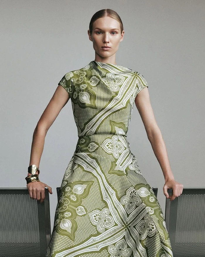 Model wearing a patterned green dress and gold cuff bracelets posing against a neutral grey studio background