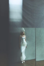 Model wearing a white dress and intricate headpiece standing against a textured wall in a muted interior space