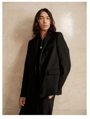 Model with long dark hair wearing a black blazer and necklace leaning against a neutral stone wall