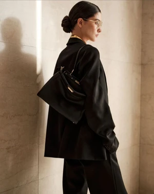 Model in a black suit holding a black handbag against a neutral stone wall in profile view.
