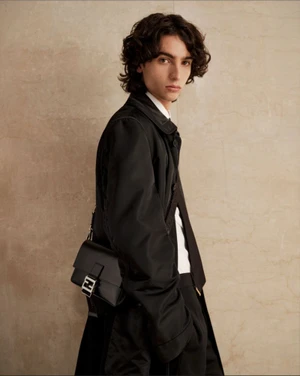 A young man with dark curly hair wearing a black coat and carrying a black handbag against a neutral wall background.