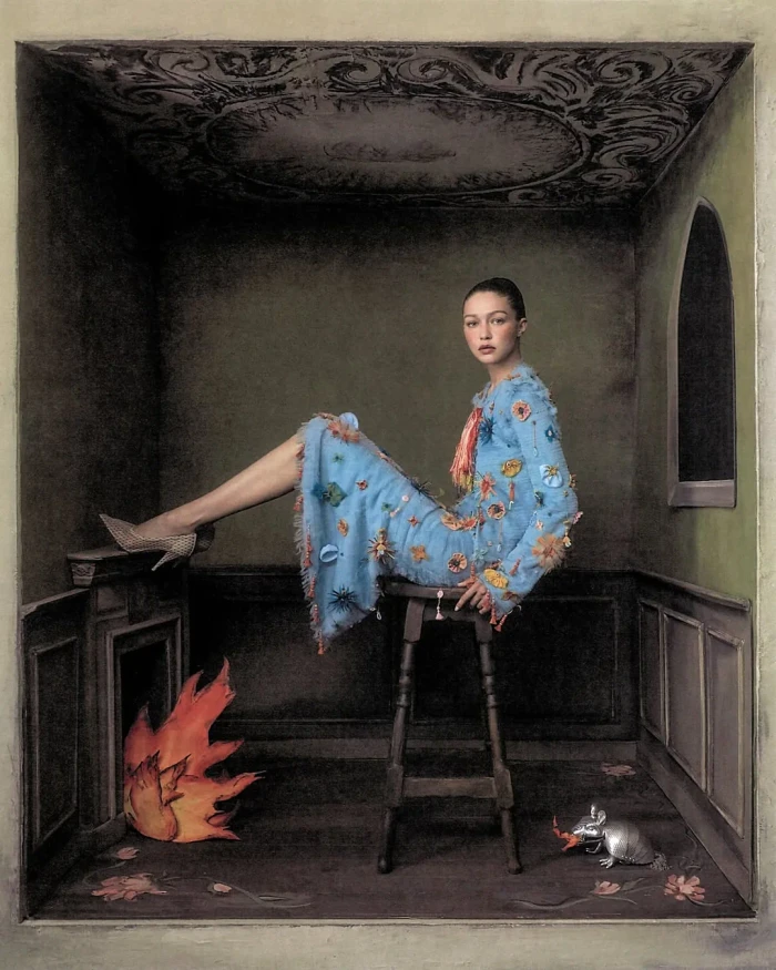 Gigi Hadid seated on a wooden stool wearing a textured blue embroidered dress inside a surreal miniature room set