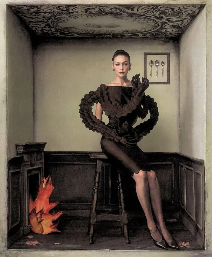 Gigi Hadid seated on a wooden stool wearing a black sculptural dress with connected gloves in a small room set for WSJ.