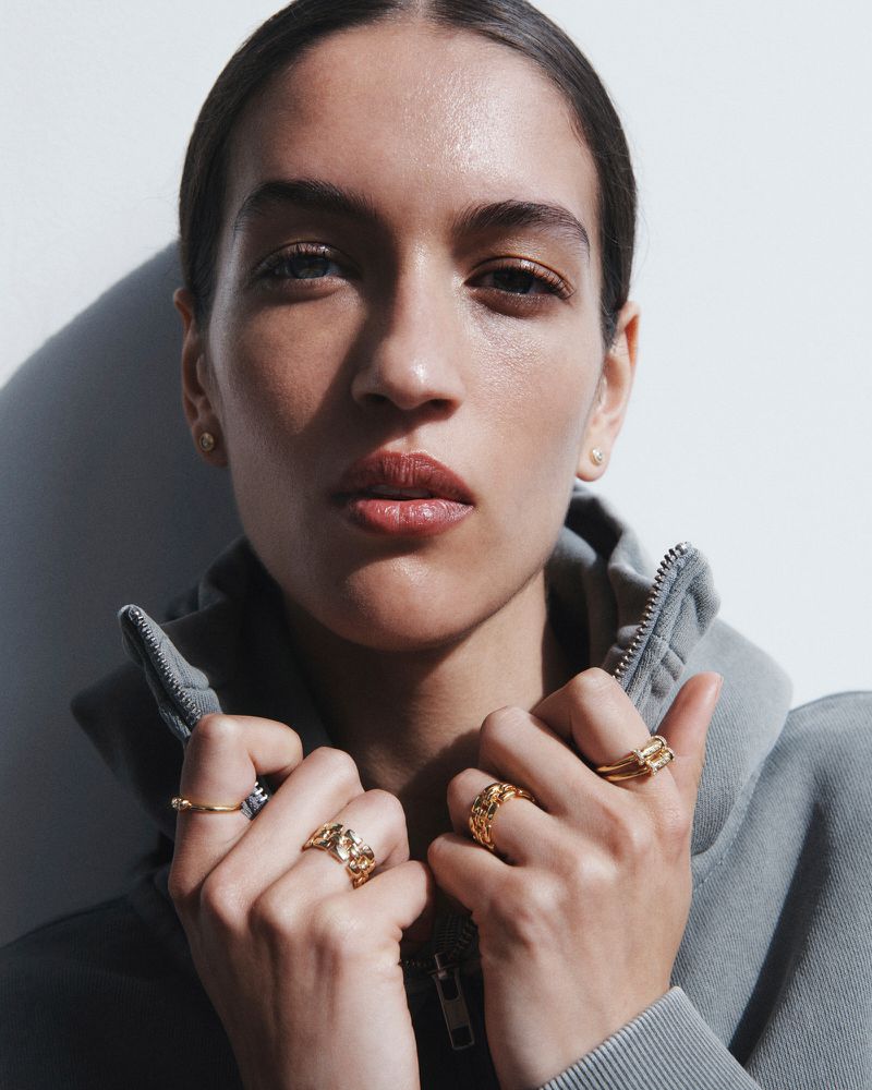 Close-up of a young adult female model wearing gold rings and a grey hooded sweatshirt against a white background in soft, diffused lighting.