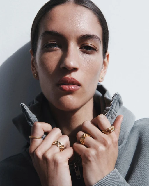 Close-up of a young adult female model wearing gold rings and a grey hooded sweatshirt against a white background in soft, diffused lighting.