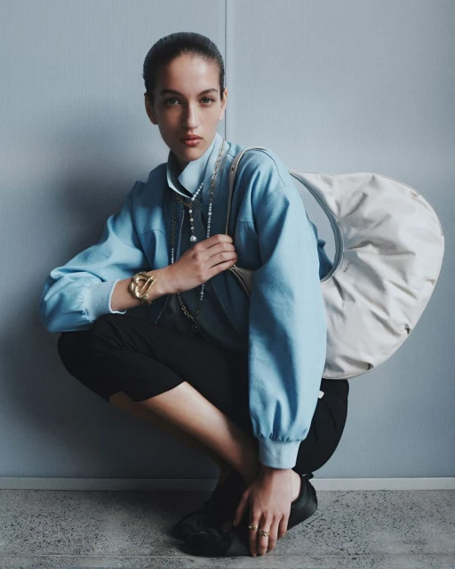 An adult female is shown crouching in a blue top and black pants accessorized with jewelry and a light-colored bag over her shoulder.