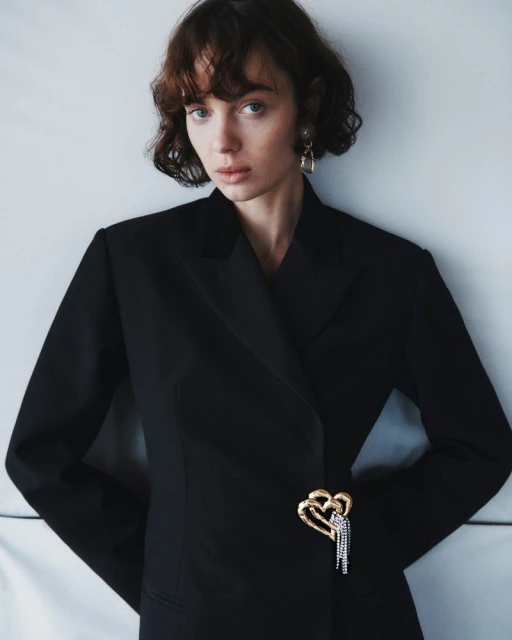Close-up of a young woman wearing a black blazer with a gold and silver heart brooch, posing indoors in a neutral setting.