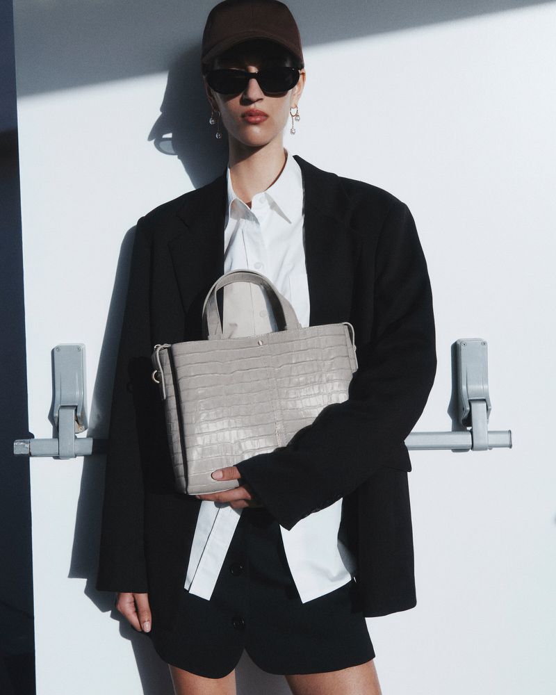 Woman in a black blazer, white shirt, and baseball cap holds a structured beige tote bag against a white door in a fashion shot.