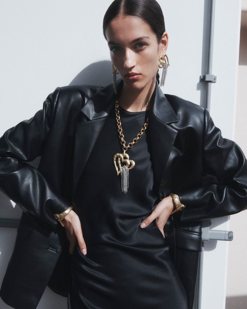 Fashionable adult woman in black leather blazer and dress accessorized with gold jewelry posing in a brightly lit indoor setting.