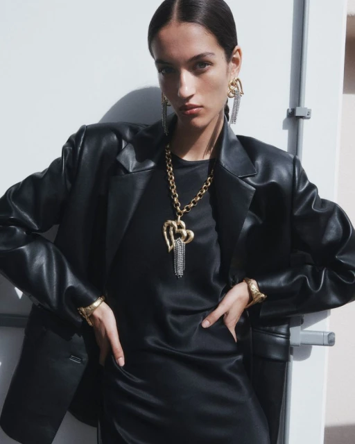 Fashionable adult woman in black leather blazer and dress accessorized with gold jewelry posing in a brightly lit indoor setting.