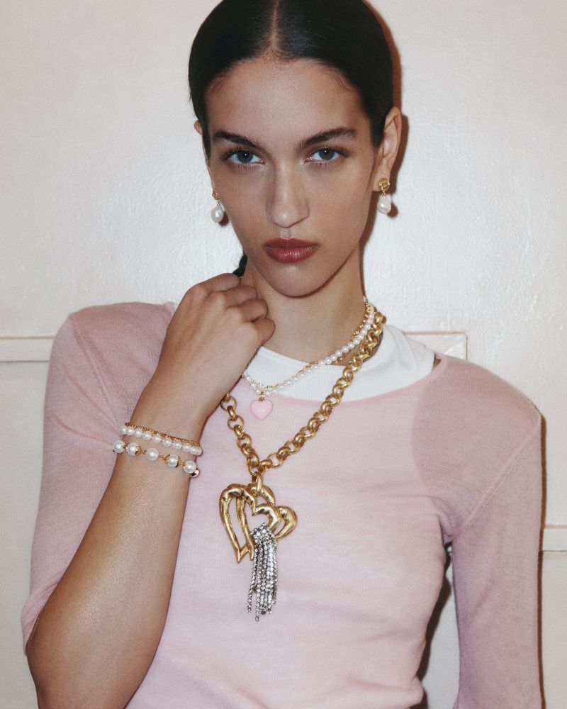A young adult female poses indoors wearing a pink top and white collar, accessorized with gold jewelry and pearl earrings.