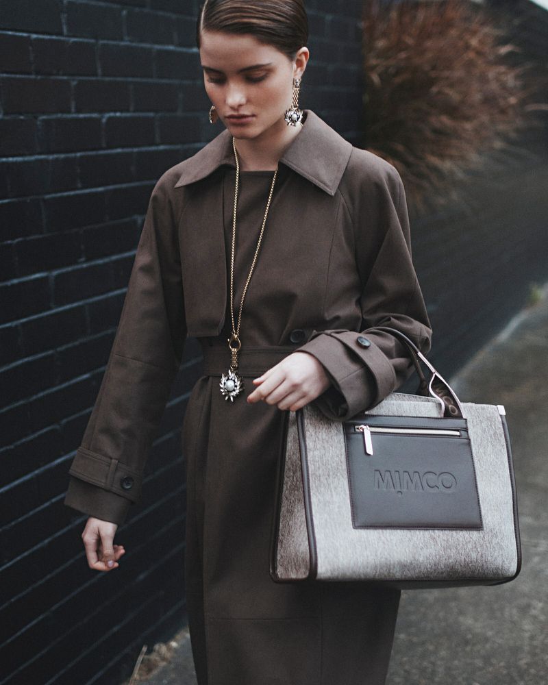 A woman in a trench coat poses with a Mimco handbag against a dark brick wall, showcasing fashion and accessory design in an outdoor setting.