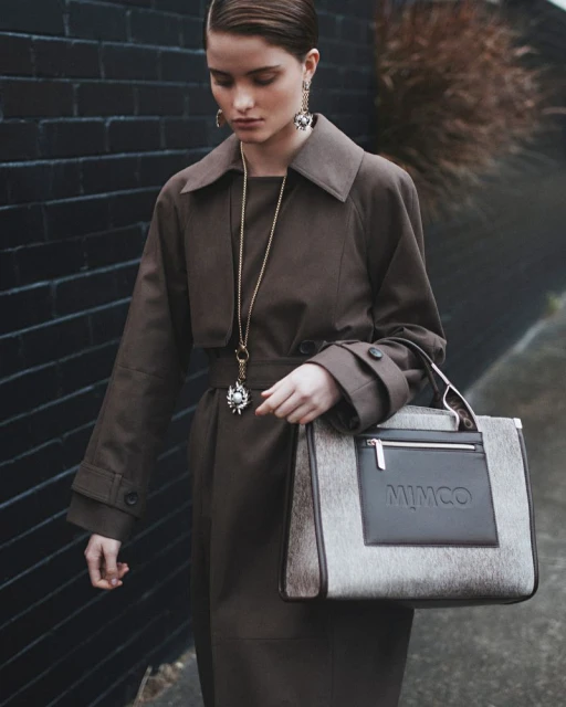 A woman in a trench coat poses with a Mimco handbag against a dark brick wall, showcasing fashion and accessory design in an outdoor setting.