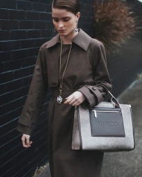 Model wearing a brown trench coat and statement pendant necklace holding a patterned handbag against a dark brick wall.