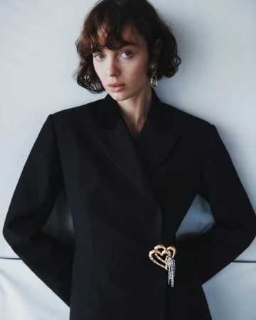 Model Lily Vander Meeden wearing a black tailored blazer adorned with a gold heart brooch against a pale studio backdrop.