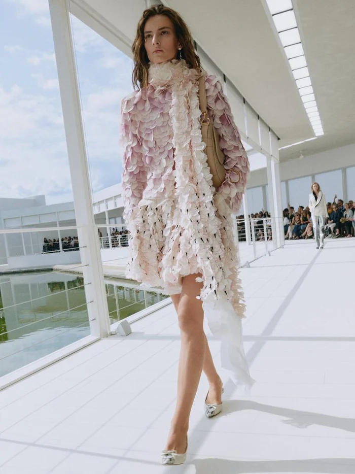 Model walking on a white runway wearing a pink textured dress and matching long scarf for Dior Summer 2027.