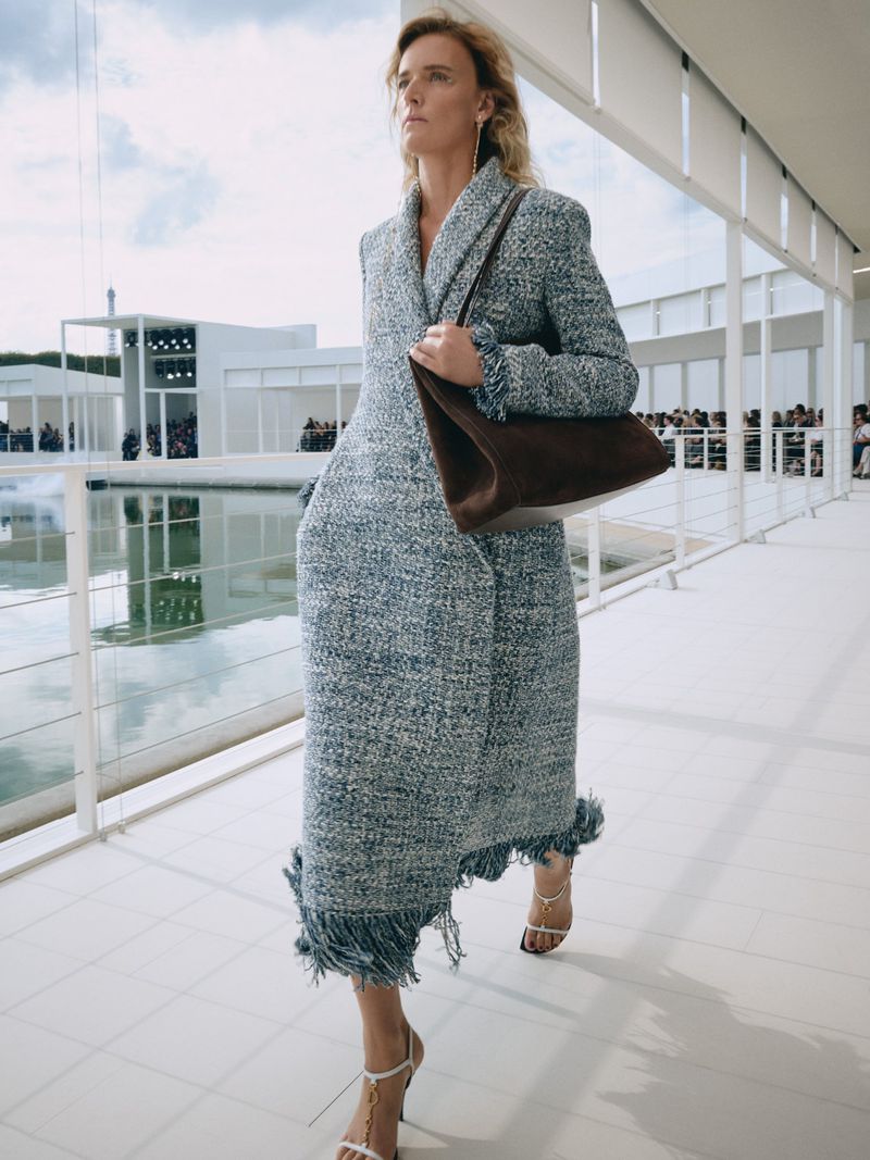 Model walking along a white pavilion walkway wearing a tweed fringe coat and holding a brown suede handbag.