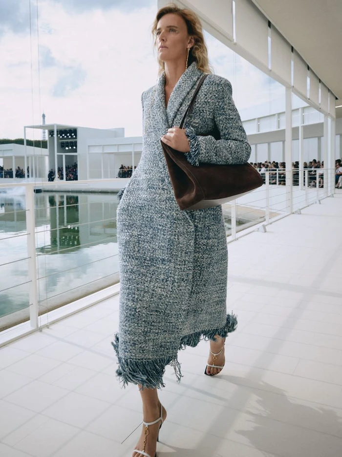 Model walking along a white pavilion walkway wearing a tweed fringe coat and holding a brown suede handbag.