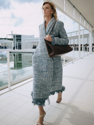 Model walking along a white pavilion walkway wearing a tweed fringe coat and holding a brown suede handbag.