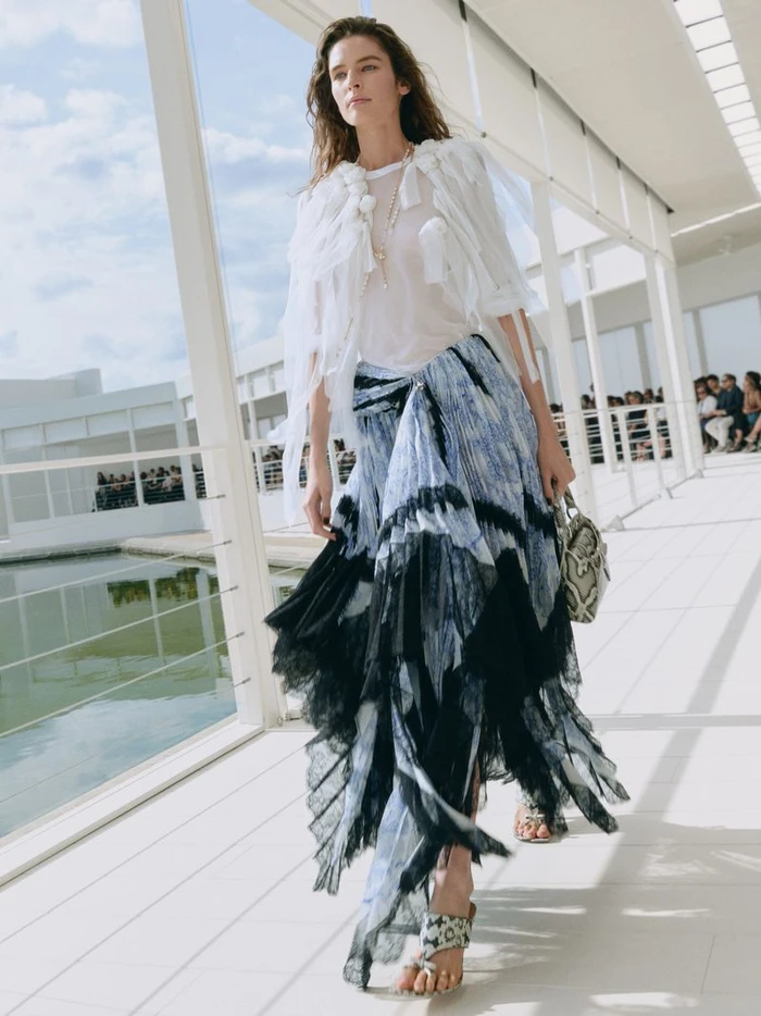 Model walking on a white terrace wearing a white sheer top and a blue patterned tiered skirt during a Dior fashion show.