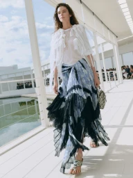 Model walking on a white terrace wearing a white sheer top and a blue patterned tiered skirt during a Dior fashion show.