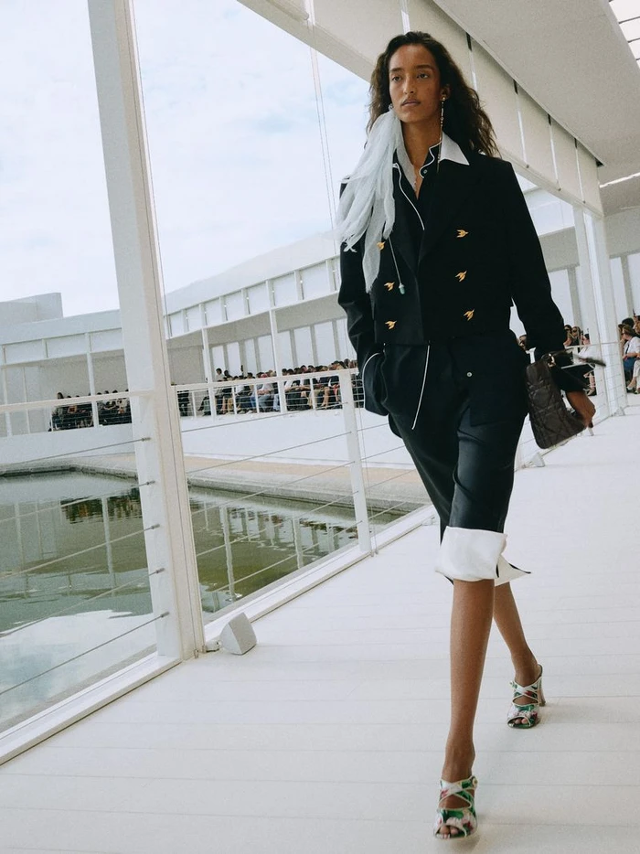 Model walking a runway in a black jacket and cropped trousers with a light scarf for Dior Summer 2027 Show.