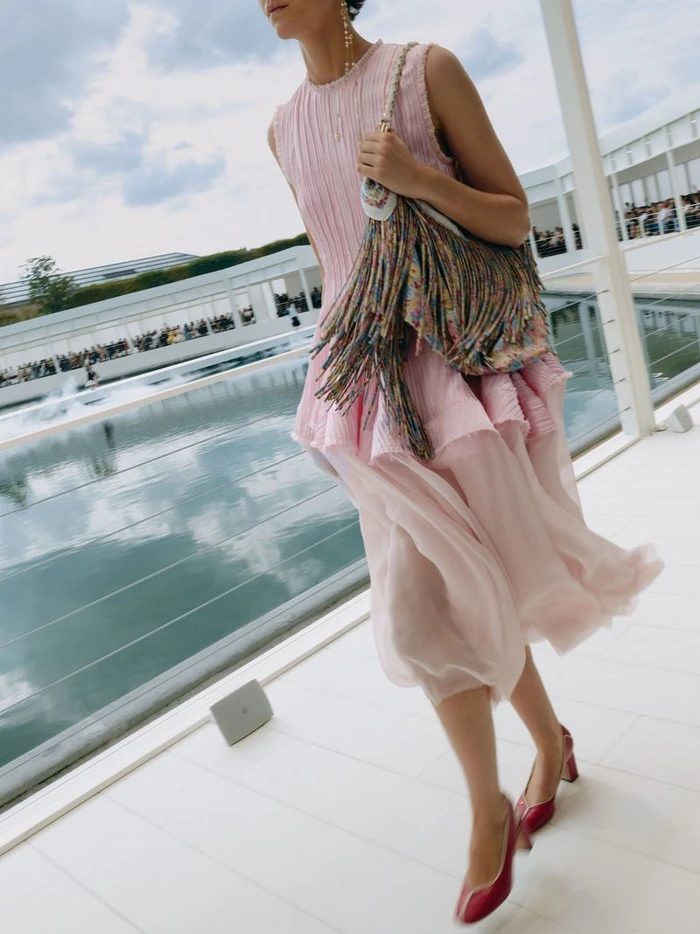 Model walking in a pleated pink dress carrying a fringe handbag outdoors near a body of water for Dior Summer 2027.