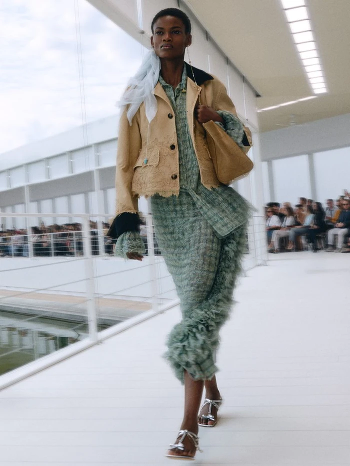 Model walking a runway wearing a textured green dress, tan jacket, and carrying a beige handbag for Dior Summer 2027.