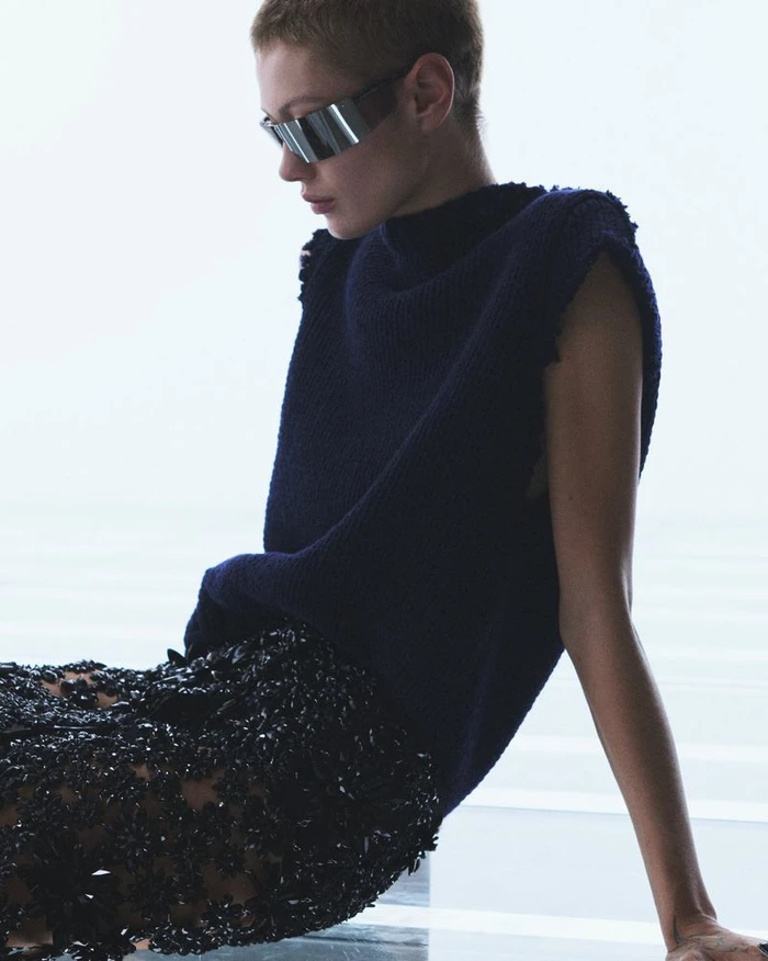 Model wearing dark futuristic sunglasses, a navy blue sleeveless top, and a heavily textured black skirt seated against a bright white background.