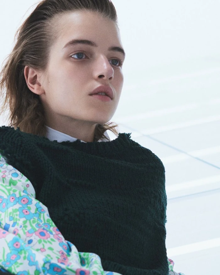 Close-up portrait of a model with slicked-back hair wearing a dark green knitted sweater and a floral patterned shirt against a plain background