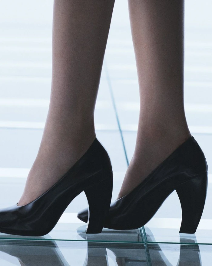 Close-up of legs wearing black heeled pumps standing on a reflective glass surface for Prada Spring/Summer 2027.