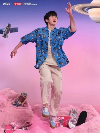 Asian male model in a patterned blue shirt jumps in front of skateboards and a planet backdrop for a Vans advertisement.