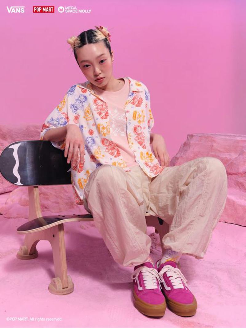 Young woman in patterned shirt sits on a skateboard bench against a pink background, wearing pink sneakers, in a studio setting.