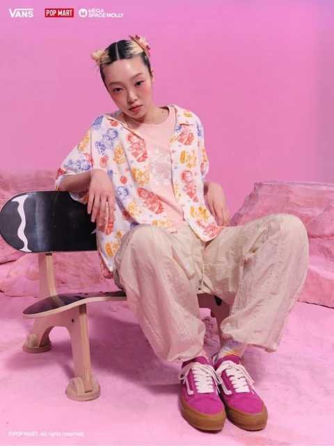 Young woman in patterned shirt sits on a skateboard bench against a pink background, wearing pink sneakers, in a studio setting.