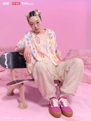 Young woman seated on a wooden chair wearing printed button-up shirt and pink sneakers against a pink studio background.