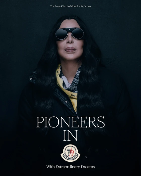 Cher wearing dark round sunglasses and a dark puffer jacket against a dark background for Moncler Re Icons