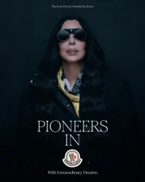 Cher wearing dark round sunglasses and a dark puffer jacket against a dark background for Moncler Re Icons