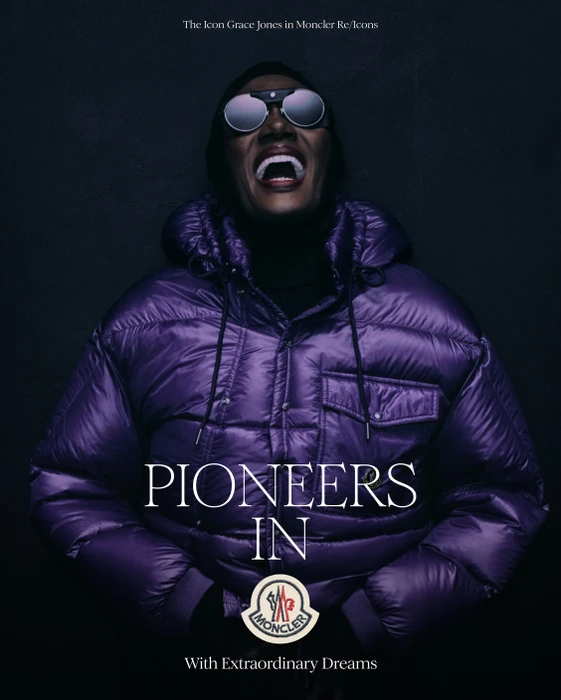 Grace Jones wearing a glossy purple puffer jacket and round sunglasses with an open mouth expression against a dark background