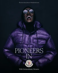 Grace Jones wearing a glossy purple puffer jacket and round sunglasses with an open mouth expression against a dark background