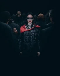 Woman in dark sunglasses, red scarf, and black puffer jacket standing amidst a blurred crowd under low key lighting.