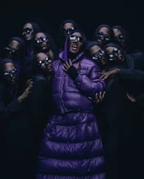 Group of black models in dark sunglasses and hooded garments surrounding a central person in a glossy purple quilted puffer jacket.