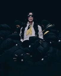 Cher wearing a white puffer jacket and yellow scarf surrounded by dark puffer jackets in a dark setting