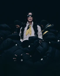 Cher wearing oversized sunglasses, a white puffer jacket, and yellow scarf, surrounded by figures in black puffer jackets
